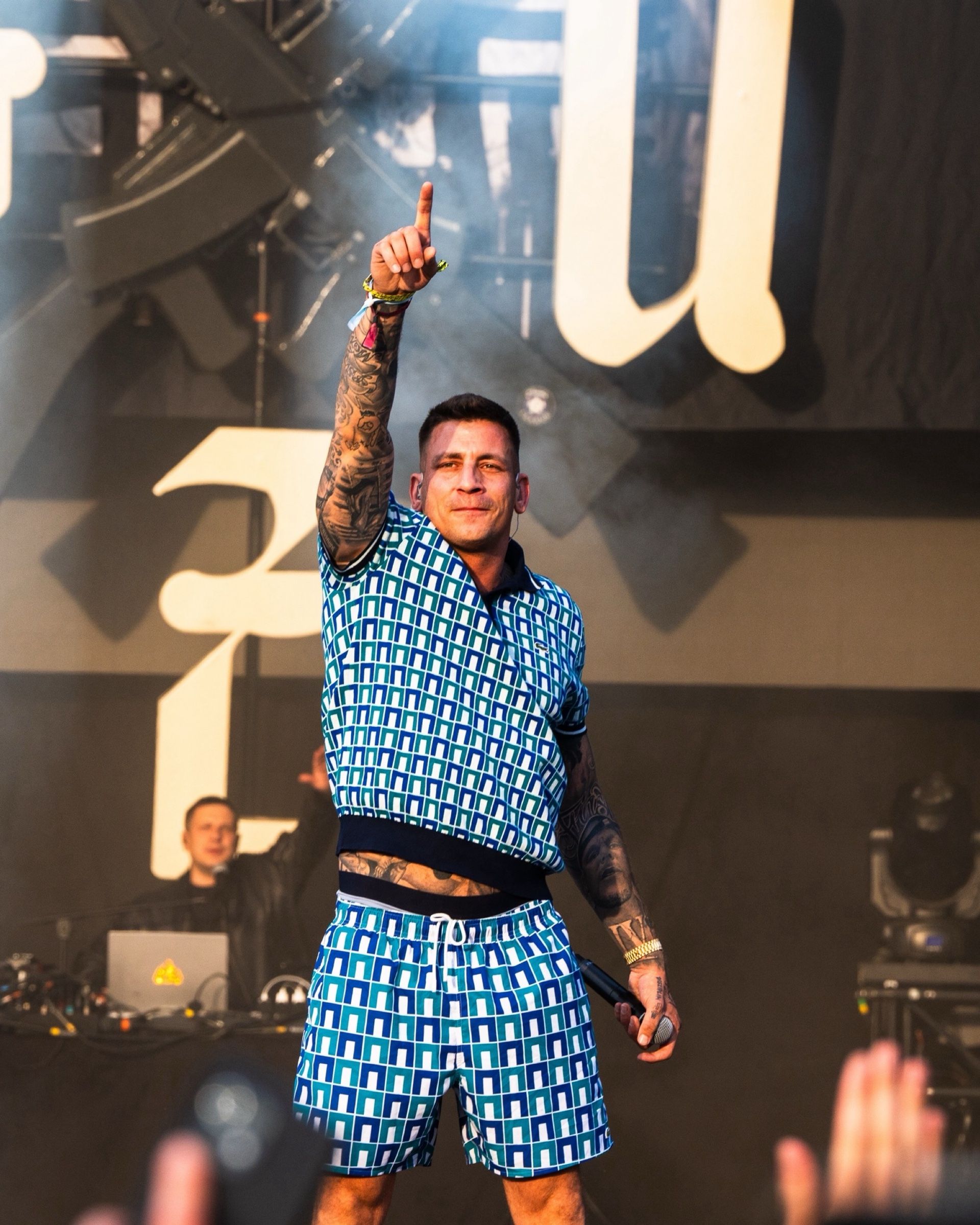 Rapper im blauen Muster-Outfit zeigt nach oben, dahinter das DJ-Setup